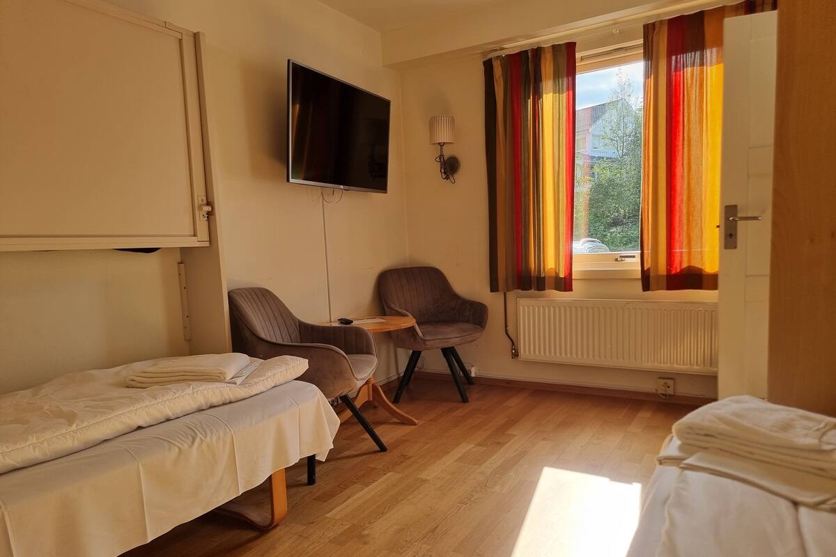 Ein Zimmer mit zwei Einzelbetten, einem an der Wand befestigten Fernseher, einem kleinen Tisch, zwei Stühlen und einem Fenster mit bunten Vorhängen.