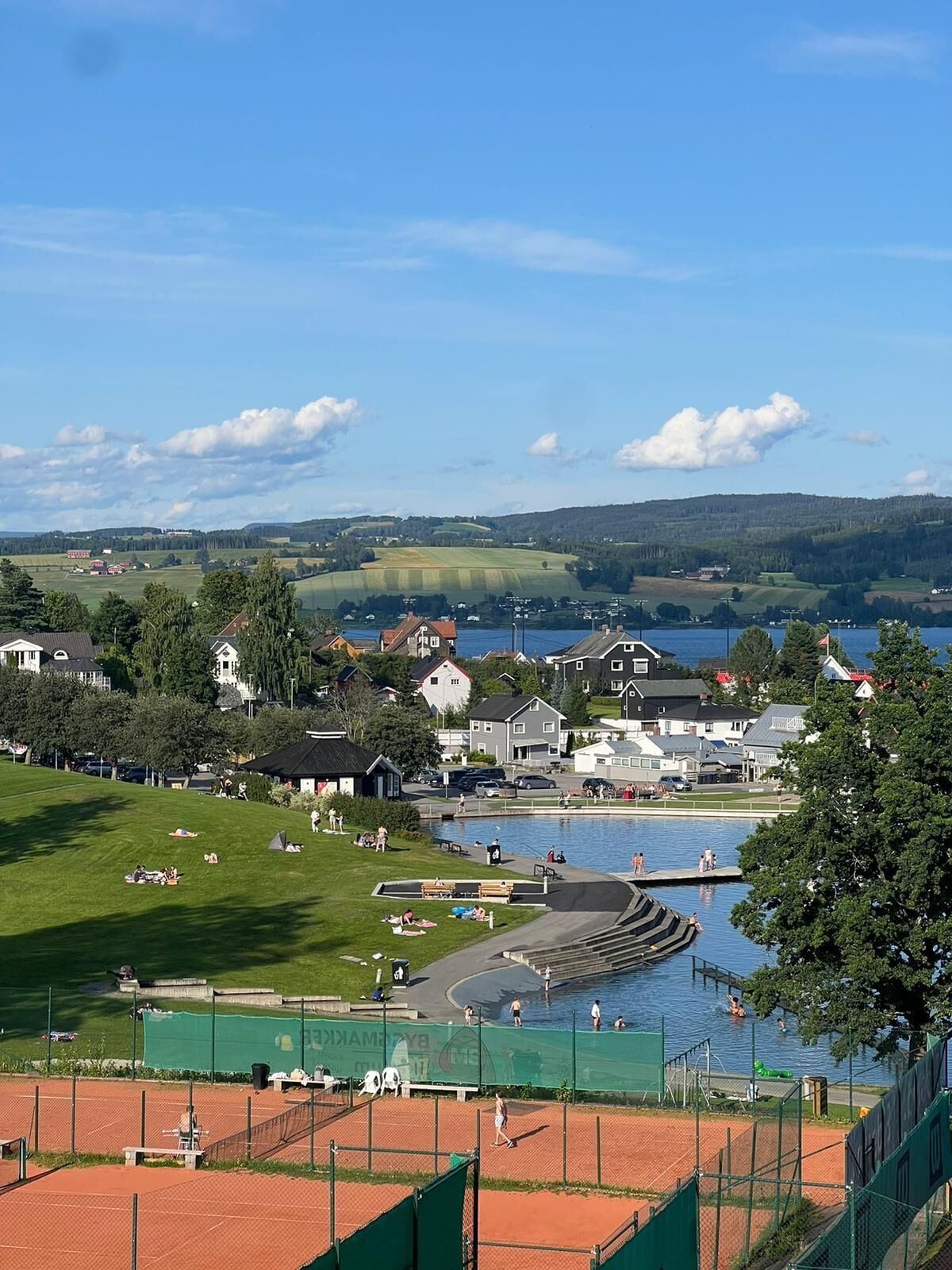 A lakeside view with a tennis court, a swimming pool, and houses in the background.