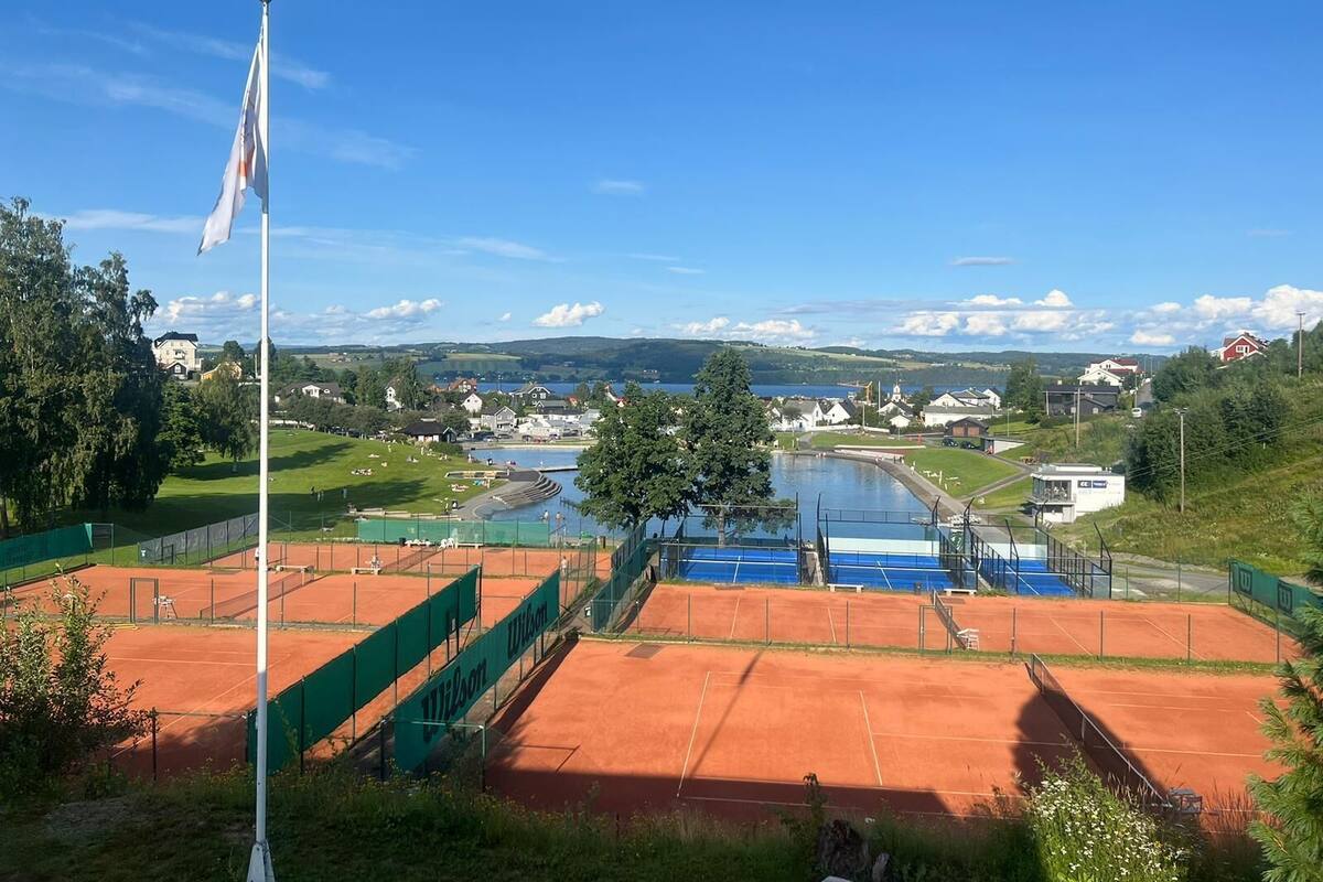 Ein Tennisplatz mit Fahnenmast und Blick auf eine Stadt und einen See im Hintergrund.