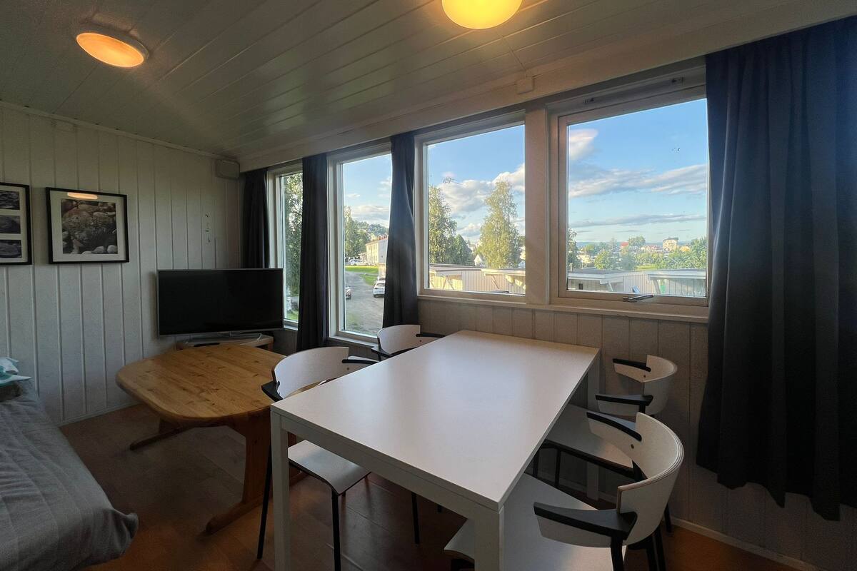 Ein Zimmer mit Holztisch und -stühlen, einem Fernseher und einem Fenster mit Vorhängen.