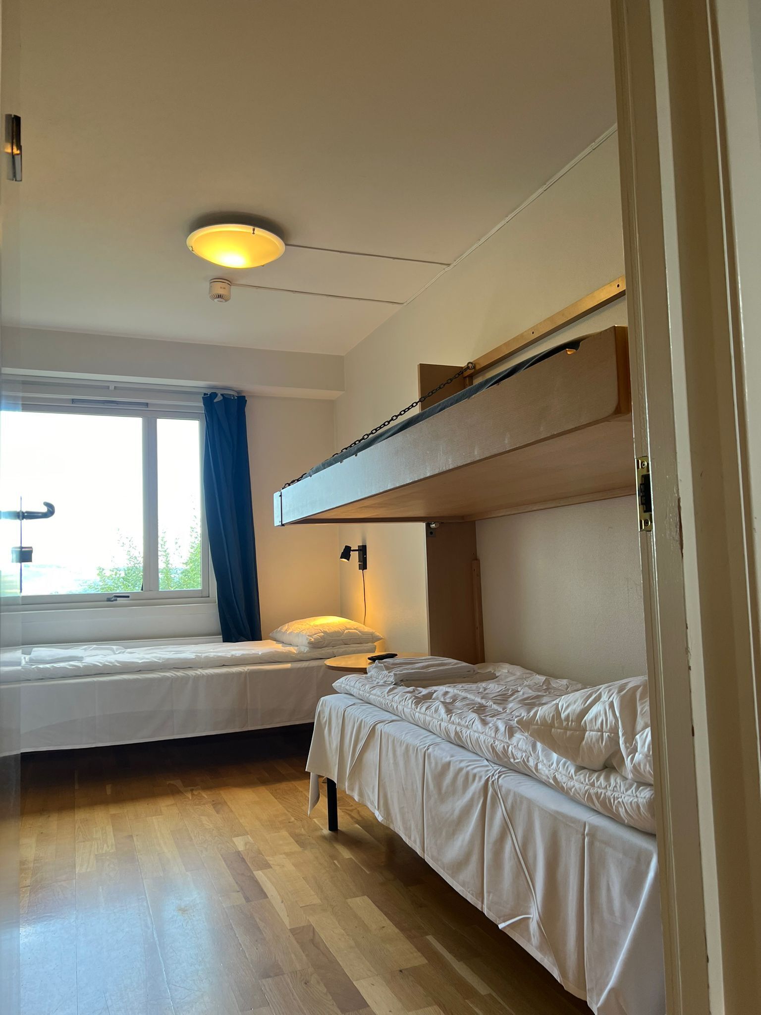 Een kamer met twee bedden, een raam met gordijnen, een plafondlamp en een houten vloer.