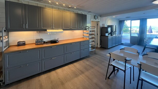 A modern kitchen with grey cabinets, a wooden countertop, and a dining area with white tables and chairs. - Hovdetun - Gjøvik Vandrerhjem - Hostel (Gjovik)