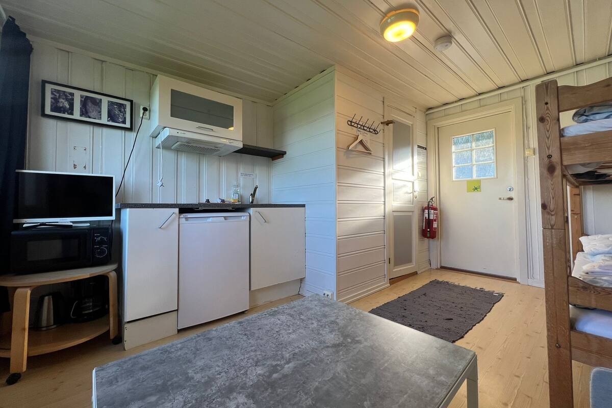 Eine kompakte Küche mit weißen Schränken, einer Mikrowelle und einem kleinen Kühlschrank. In der Mitte steht ein Tisch, daneben eine hölzerne Bank und eine Tür mit Fenster.