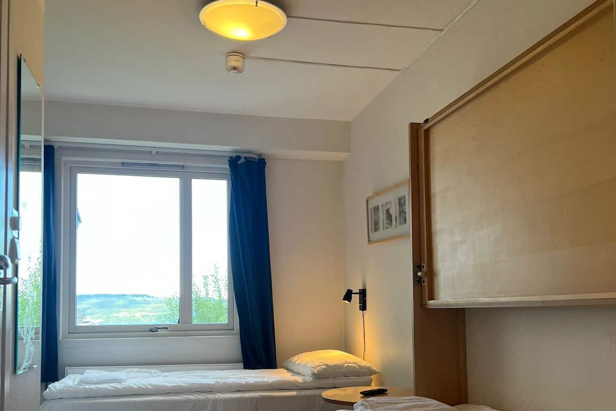 Ein Zimmer mit zwei Betten, einem Fenster mit blauen Vorhängen, einem Nachttisch mit Lampe und einer Deckenleuchte.