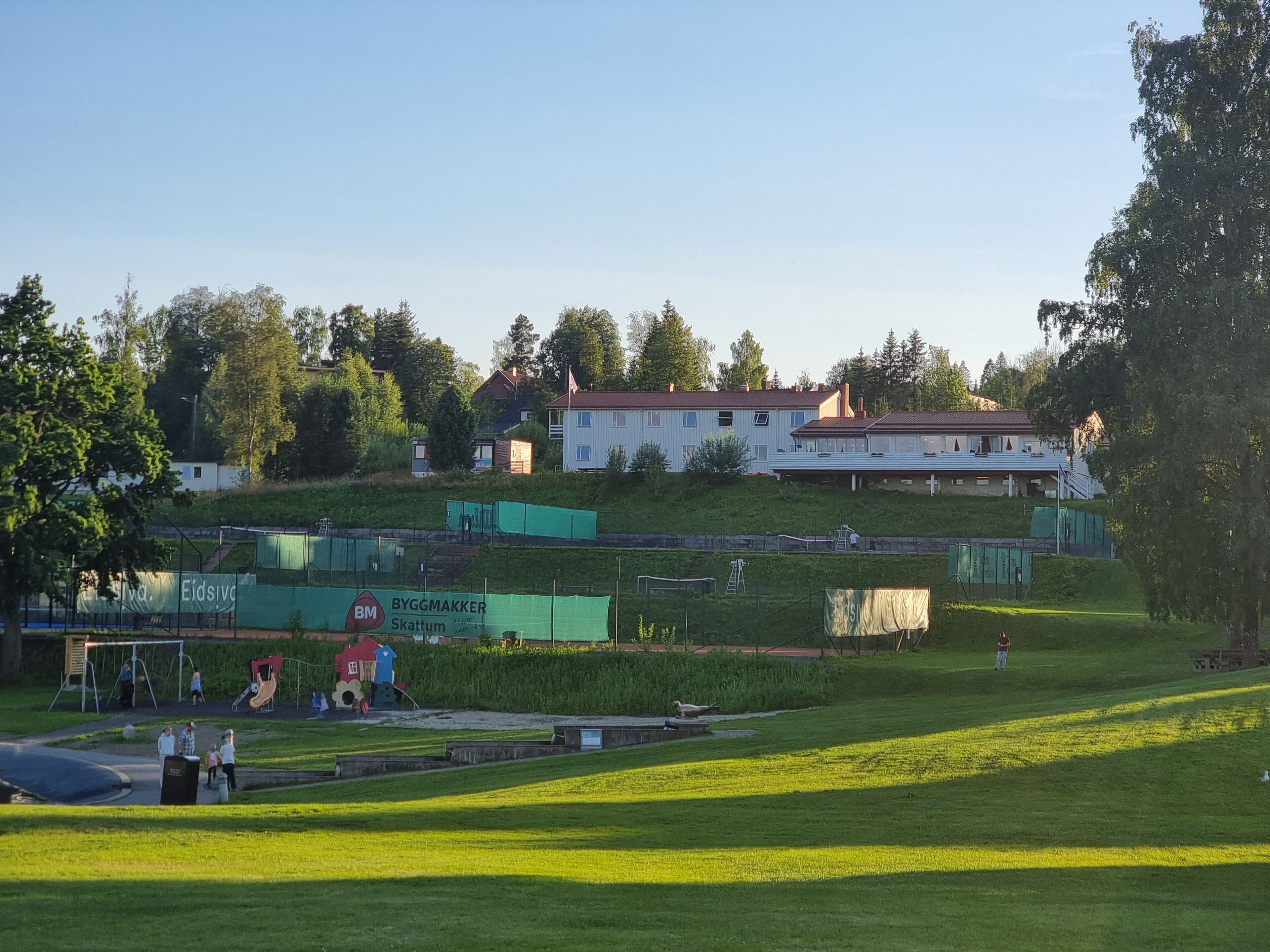 Un parque con césped, juegos infantiles, un pequeño estanque y un edificio al fondo.