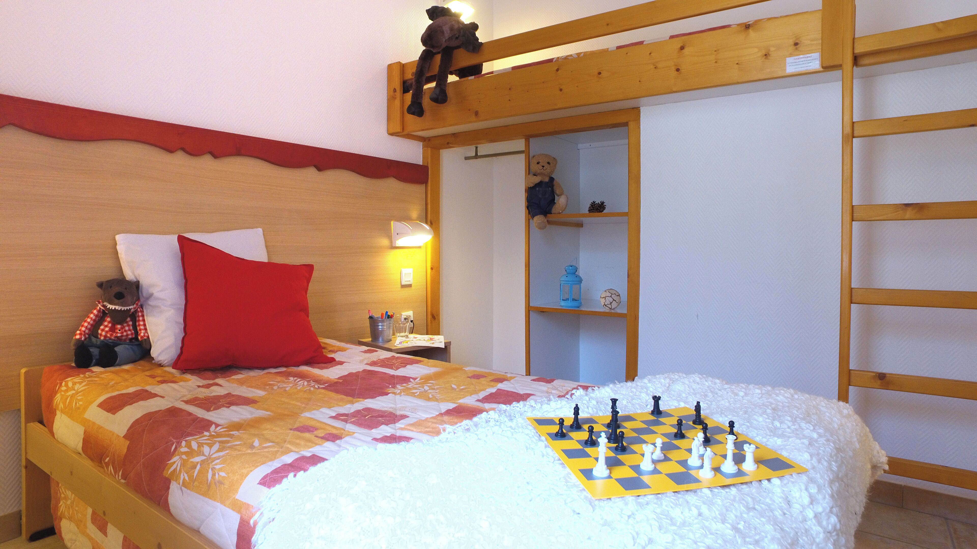 Une chambre à coucher avec un lit, un oreiller rouge, un ours en peluche, un jeu d’échecs et un lit en mezzanine.
