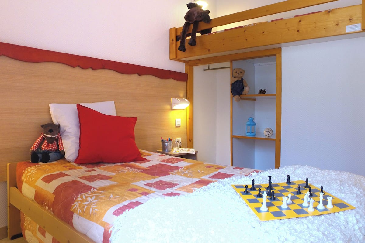 Une chambre à coucher avec un lit, un oreiller rouge, un ours en peluche, un jeu d’échecs et une échelle en bois.