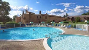 Une piscine entourée de chaises longues et de parasols, elle-même bordée d’un bâtiment en briques et d’une zone délimitée par une clôture.