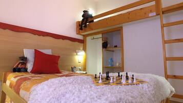 Une chambre confortable avec un lit, un oreiller rouge, un ours en peluche, une étagère avec un ours en peluche et un jeu d’échecs.