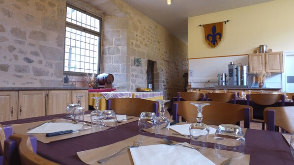 A dining area with stone walls, a wooden table set for six, and a shield emblem on the wall.
