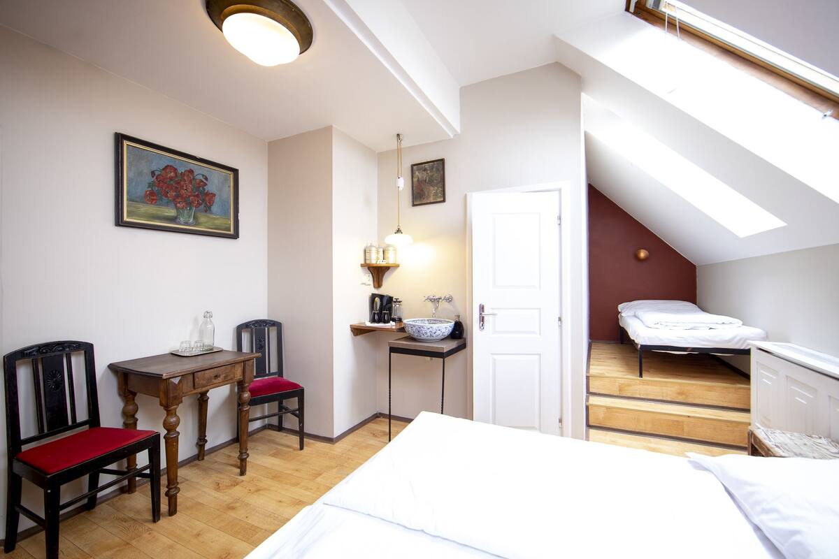 A compact room with a bed, a desk, a chair, a small table with a red cushion, a wall-mounted shelf with kitchen items, a painting, and a staircase leading to an upper level.