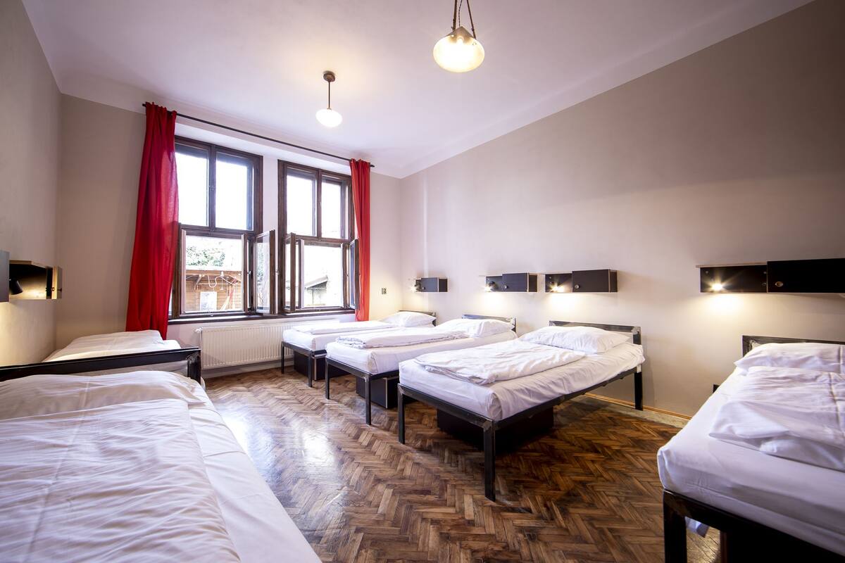 A hotel room with three beds, a large window with red curtains, and wall-mounted lights.