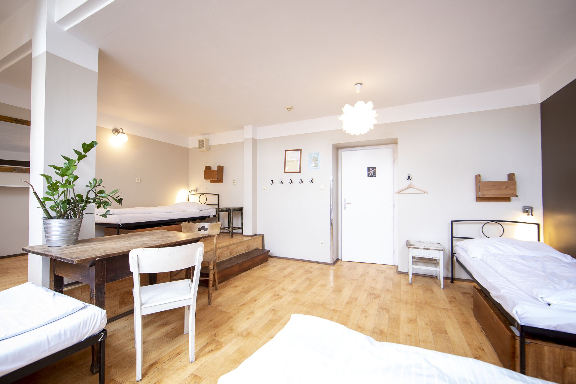 A compact hotel room with two single beds, a wooden desk, a chair, a small table, and a potted plant.