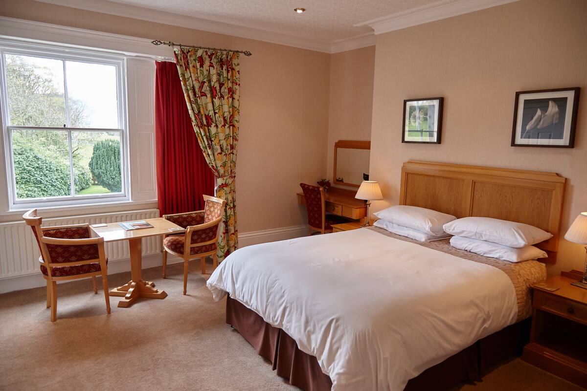 A hotel room with a large bed, a desk with a chair, a window with curtains, and a nightstand with a lamp.