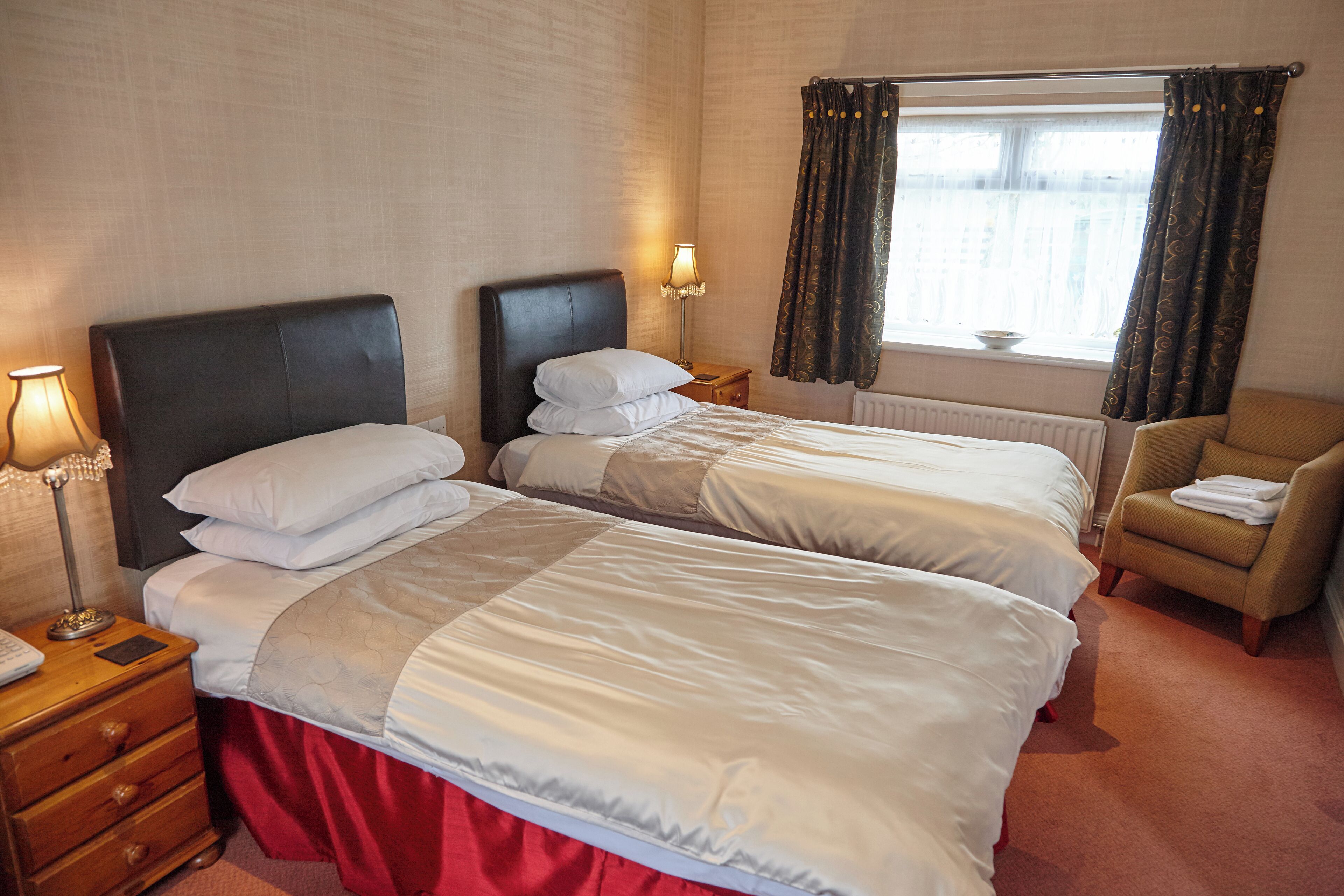 Habitación de hotel con dos camas, una mesita de noche, una lámpara, una silla y un ventanal con cortinas.