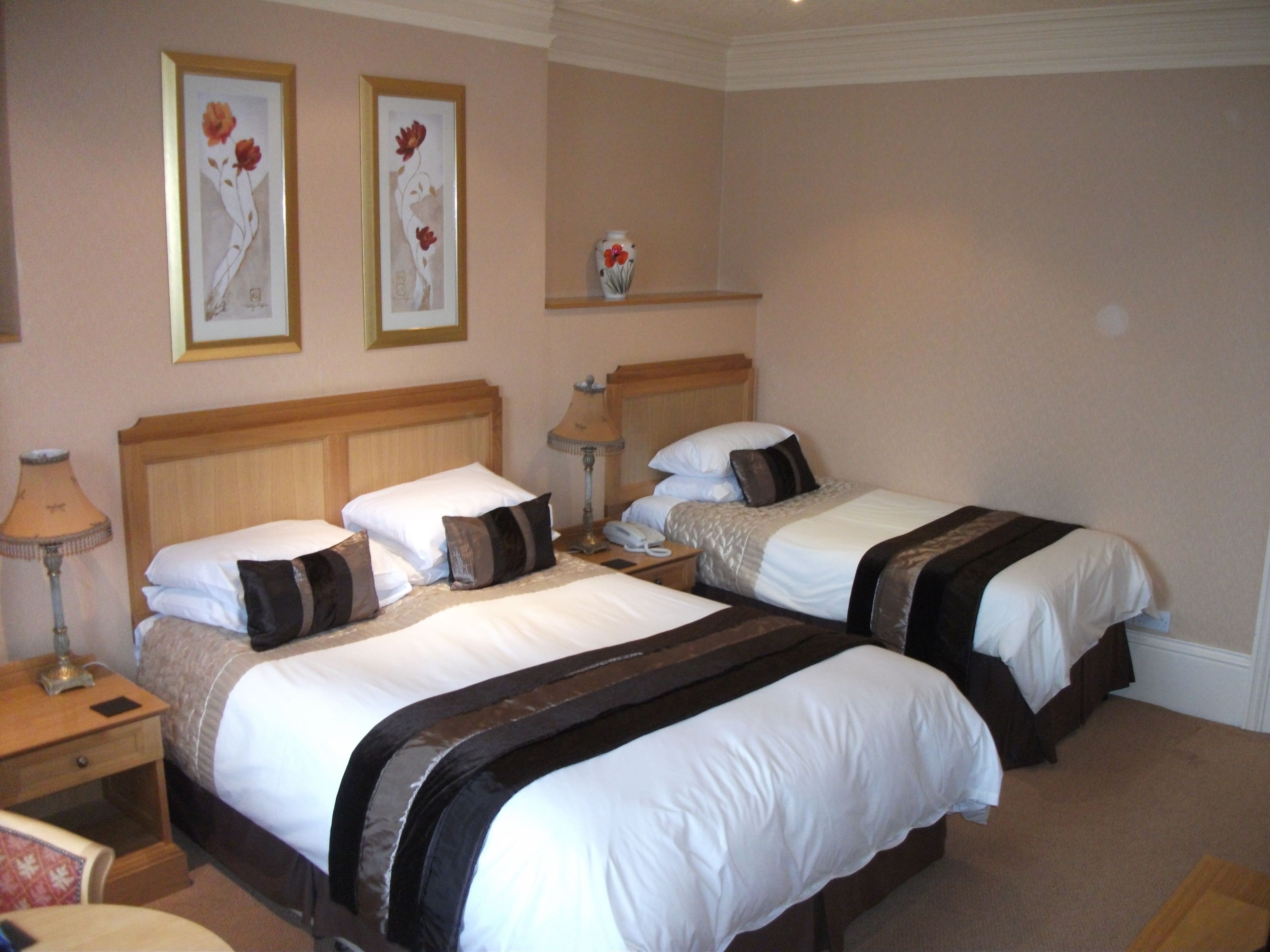A hotel room with two beds, each with white bedding and brown accents, a wooden headboard, bedside tables with lamps, and framed floral artwork on the wall.