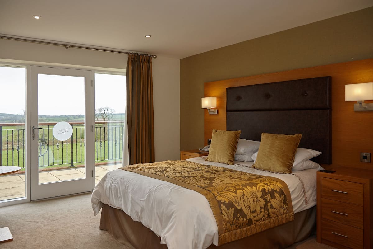 A bedroom with a large bed, two bedside lamps, a wooden headboard, and a view of a balcony with a lawn.