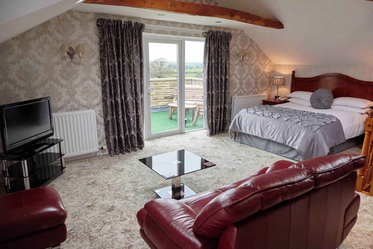 A bedroom with a bed, a TV, a red leather sofa, a glass coffee table, and a view of the outdoors.