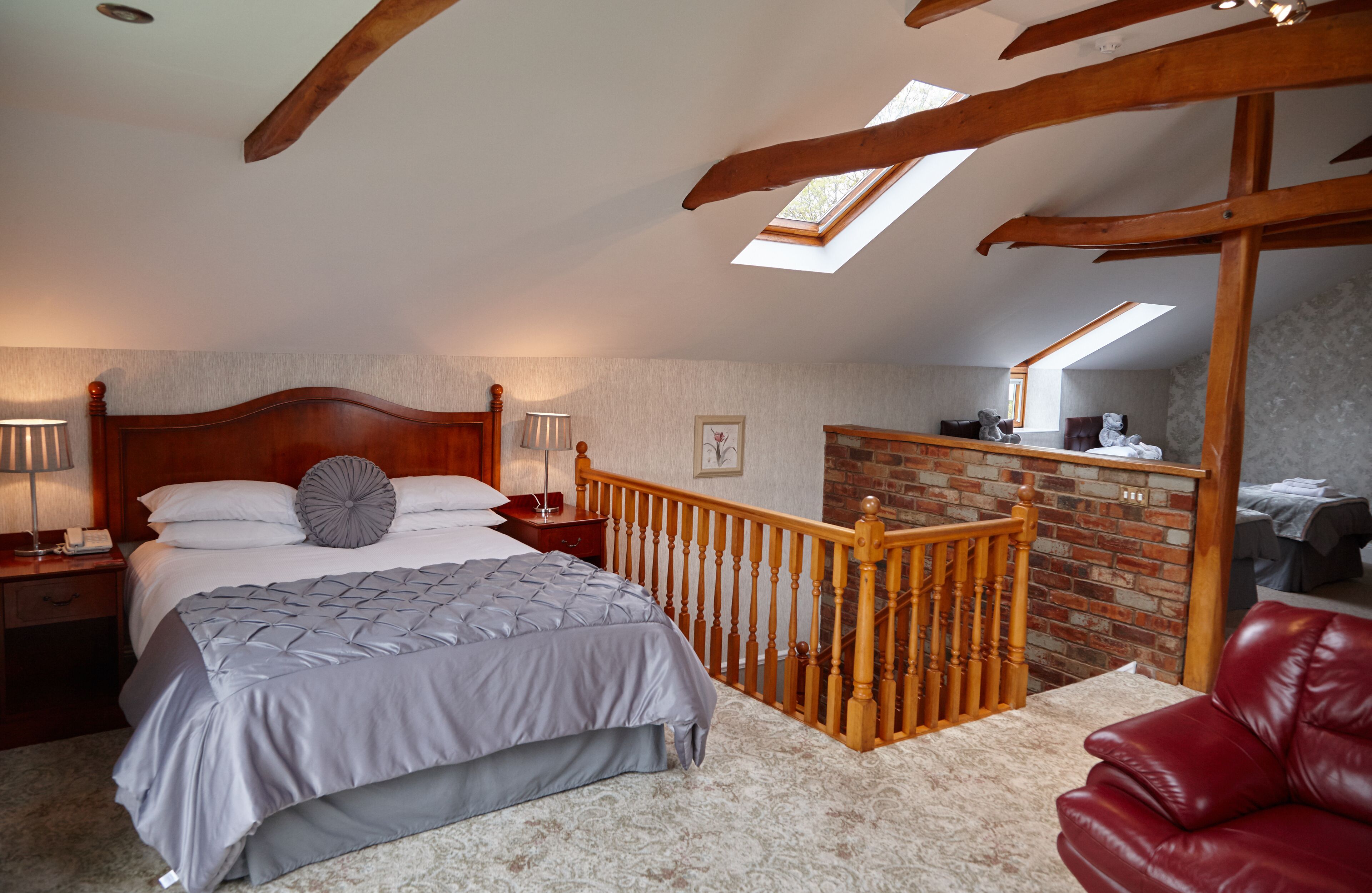 A bedroom with a large bed, wooden headboard, a nightstand, a lamp, a red leather chair, and a skylight.
