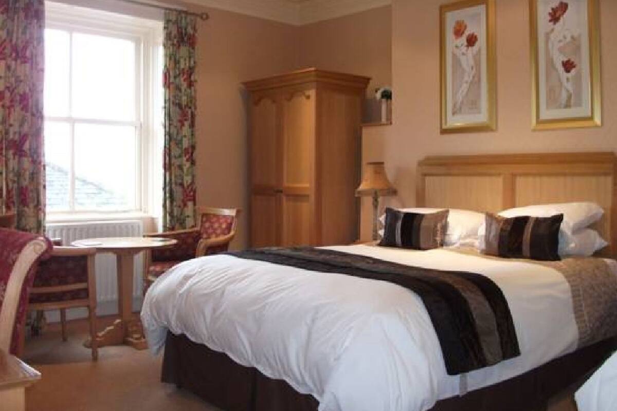A hotel room with a large bed, a wooden wardrobe, a desk with a chair, and a window with curtains.