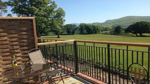 A balcony with a table and chairs, a bottle of wine, and a view of a grassy landscape with trees and hills. - Hundith Hill Hotel (Cockermouth)