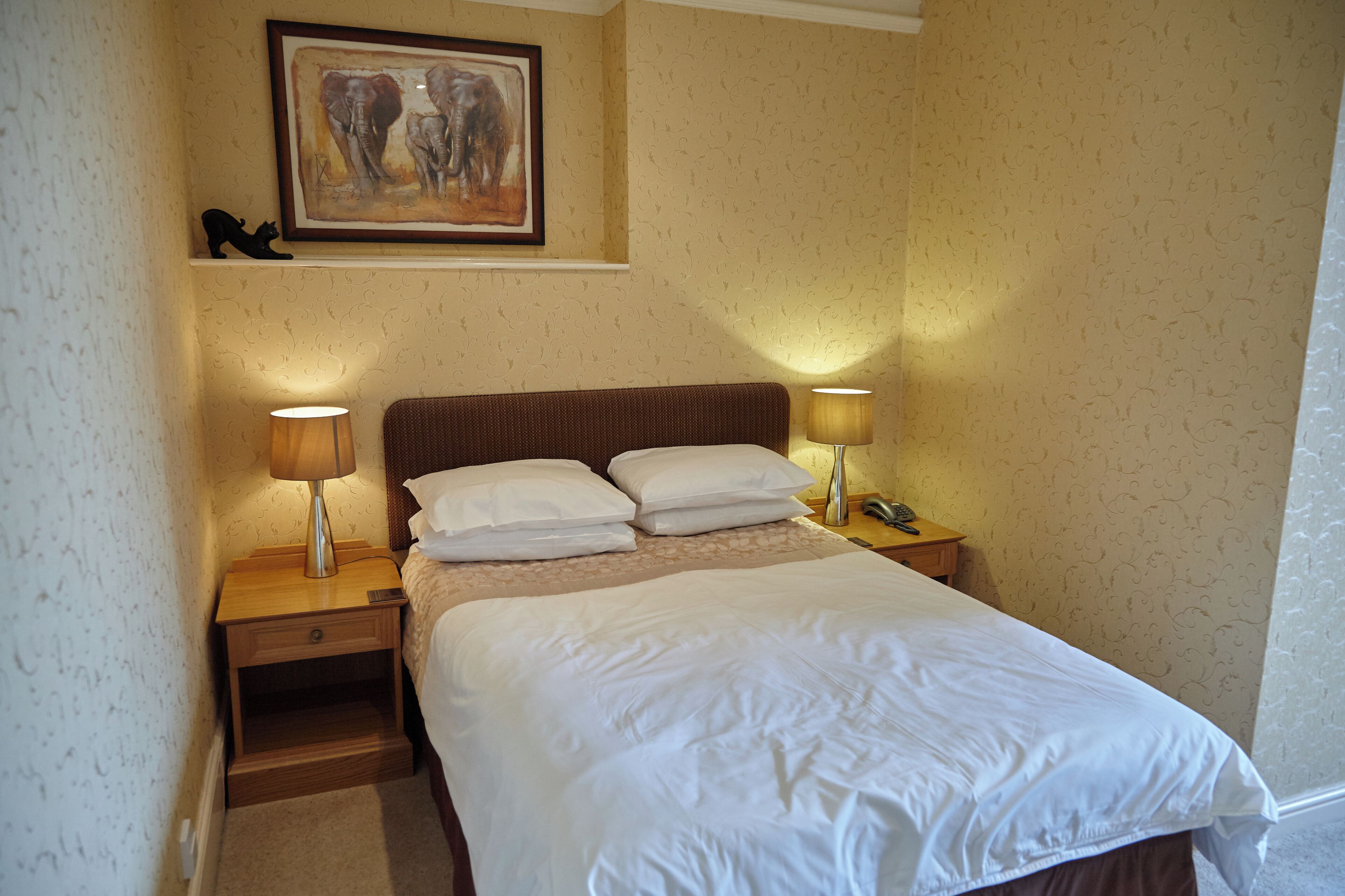A neatly made bed with white linens, two bedside lamps, a nightstand with a phone, and a framed artwork above the bed.