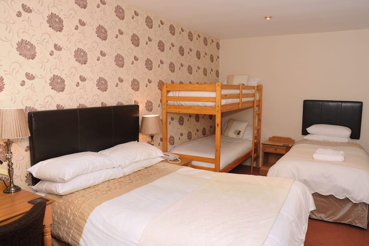 A hotel room with two beds, one with a bunk bed and the other with a single bed. There is a bedside table with a lamp and a phone. The room has floral wallpaper and a wooden headboard.
