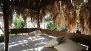 Un letto di legno con biancheria e cuscini bianchi, situato in un’area esterna coperta con tetto di paglia e palme sullo sfondo.