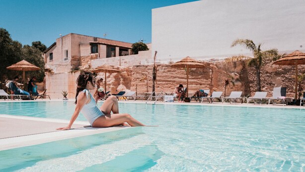Area piscina con una persona seduta sul bordo, sedie a sdraio, ombrelloni e un edificio sullo sfondo.