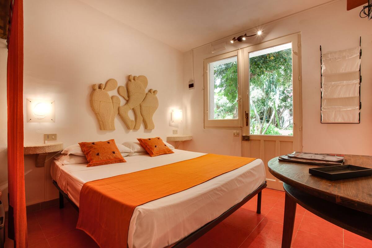 A bedroom with a bed, orange bedding, a nightstand, a wall-mounted lamp, and a window with a view of trees.