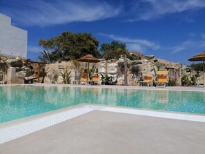 A clear swimming pool with lounge chairs and umbrellas, surrounded by rocky terrain and vegetation. - L'Oasi Villaggio Albergo (Favignana)