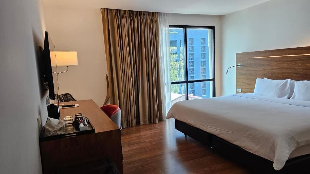 Ein Hotelzimmer mit einem großen Bett, einem Schreibtisch mit Lampe, einem Fernseher und einem Fenster mit Vorhängen.