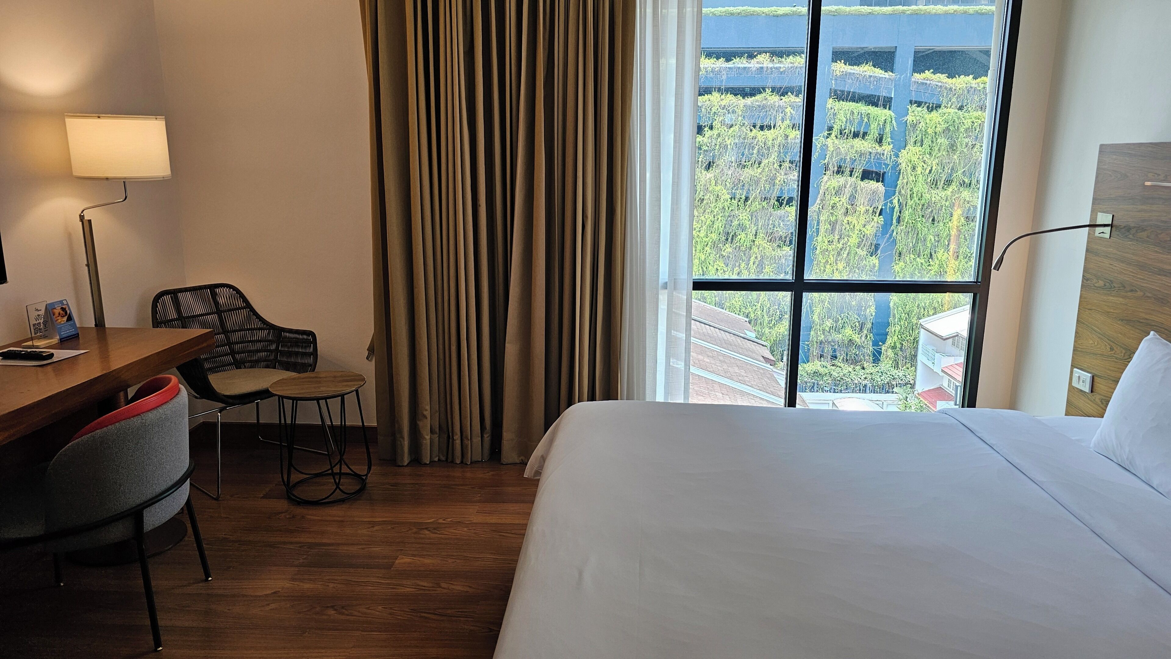 A hotel room with a bed, a desk with a chair, a lamp, and a view of greenery through the window.