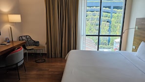 Et hotelværelse med en seng, et skrivebord med stol, en lampe og udsigt til grønne områder gennem vinduet.