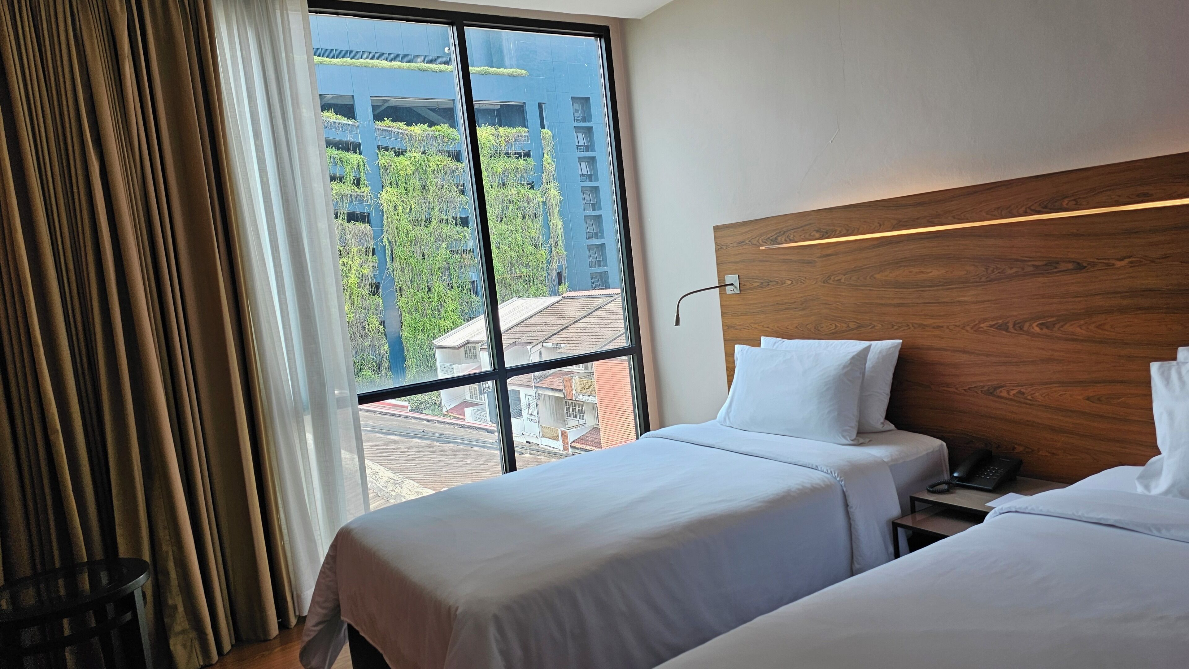A hotel room with two beds, a wooden headboard, a nightstand, a telephone, and a large window with a view of buildings and trees.