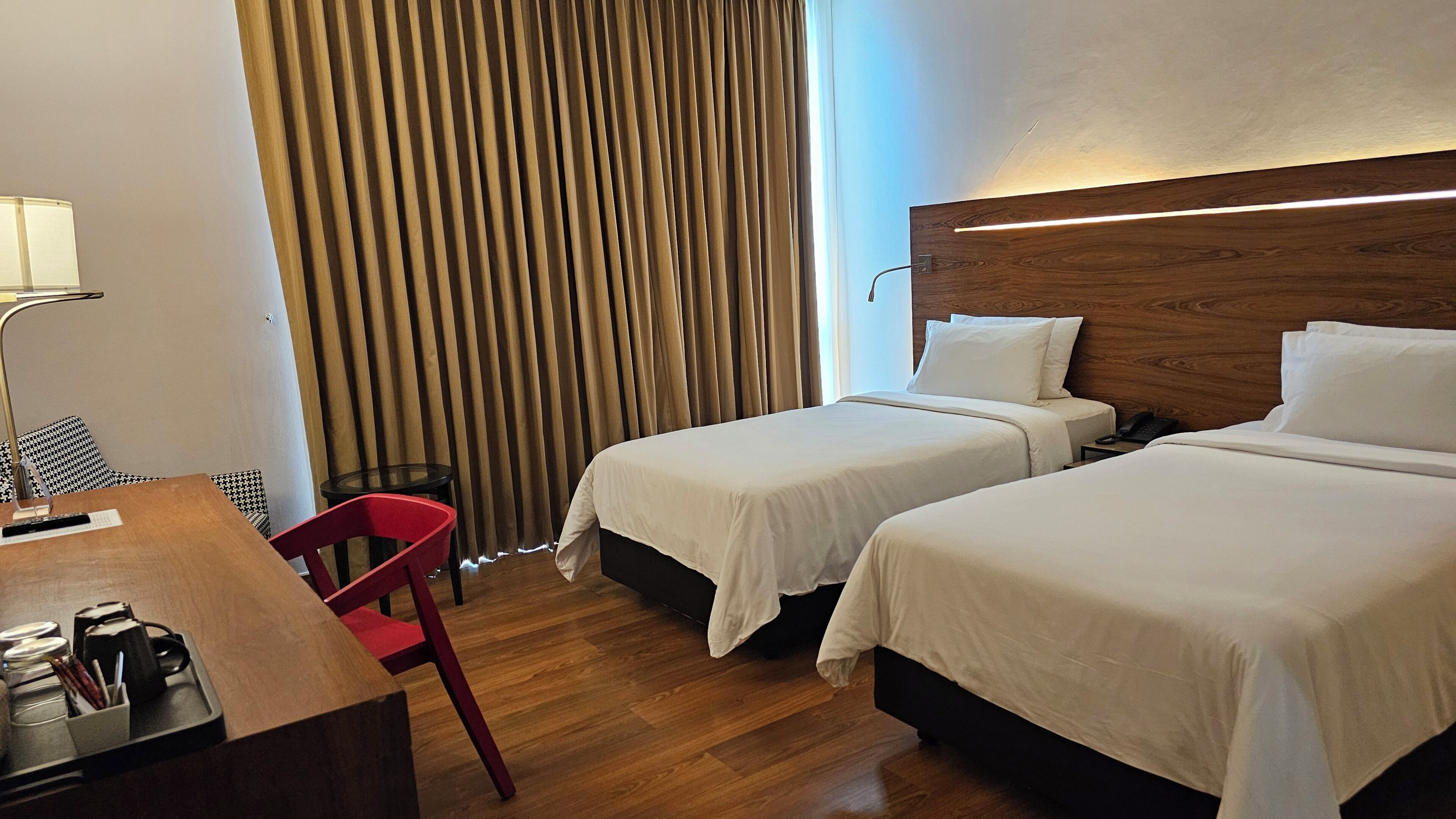 A hotel room with two beds, a desk with a lamp, a chair, and a curtain.