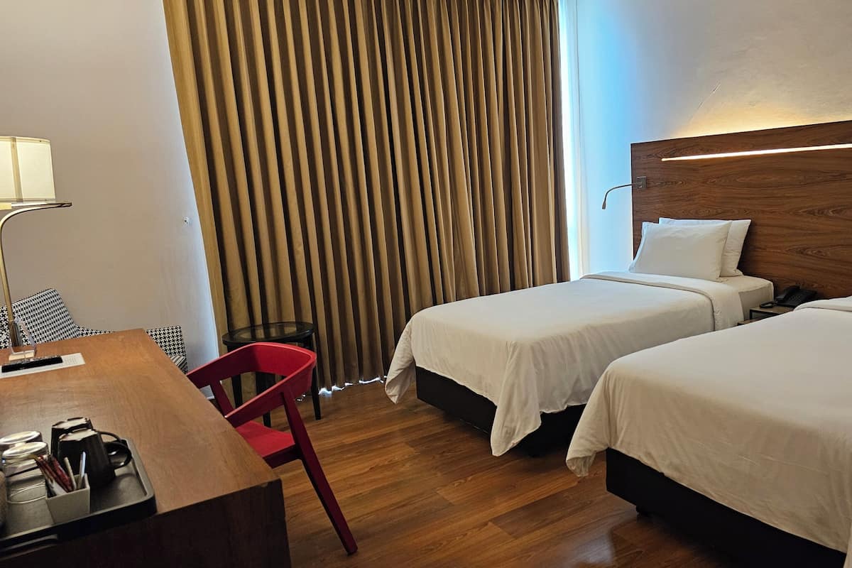 A hotel room with two beds, a desk with a lamp, a chair, and a curtain.