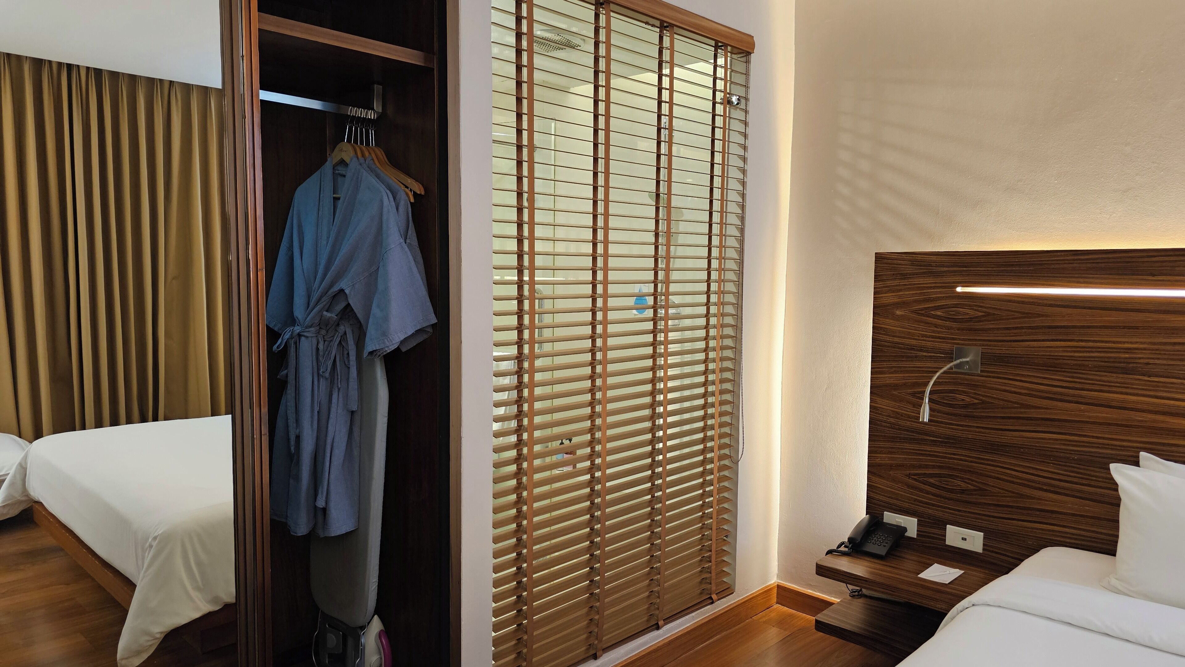 Quarto de hotel com guarda-roupa de madeira, uma cama, mesa de cabeceira com telefone e janela com persianas.