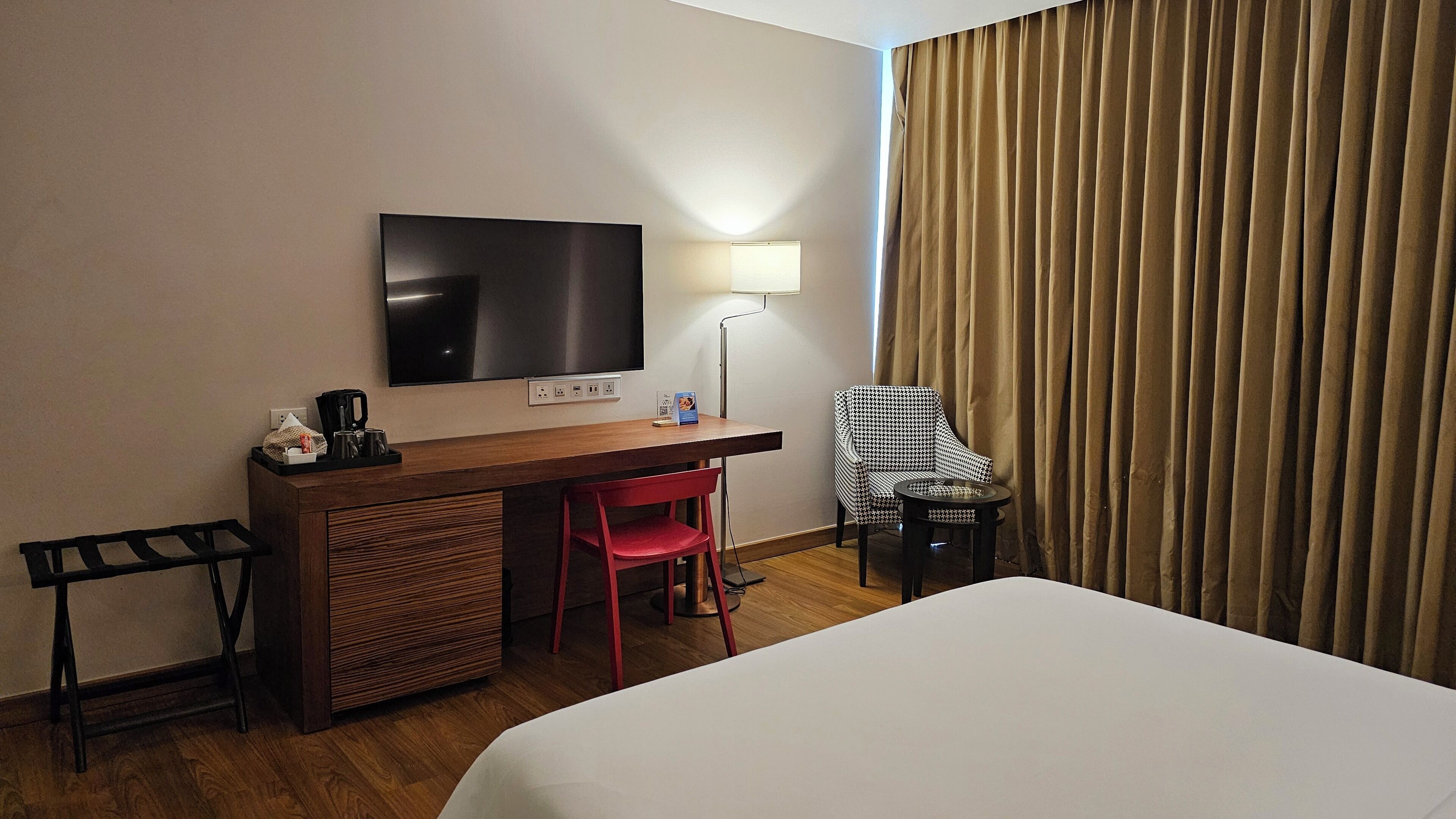 A hotel room with a bed, a desk with a chair, a television, a lamp, and a window with curtains.