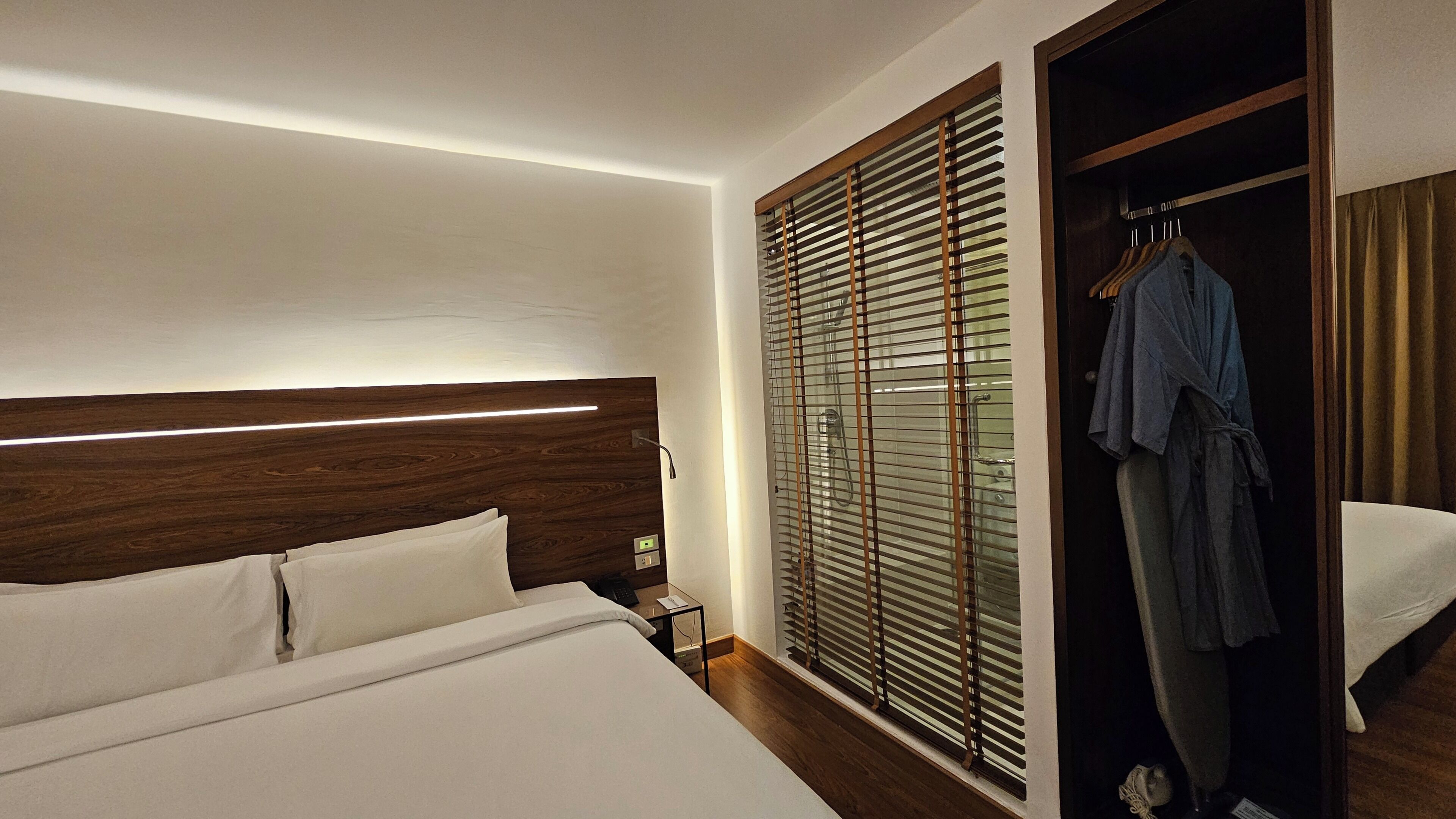A hotel room with a bed, a nightstand, a robe hanging on a door, and a window with blinds.