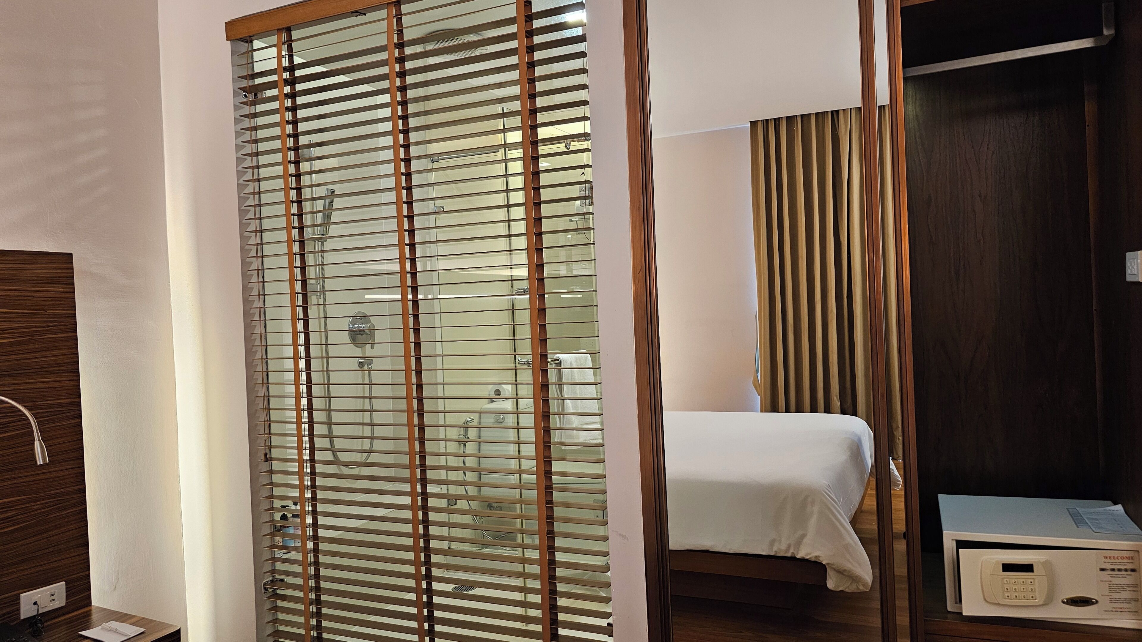 Quarto de hotel com cama, guarda-roupa de madeira, divisória de vidro e painel de controle.