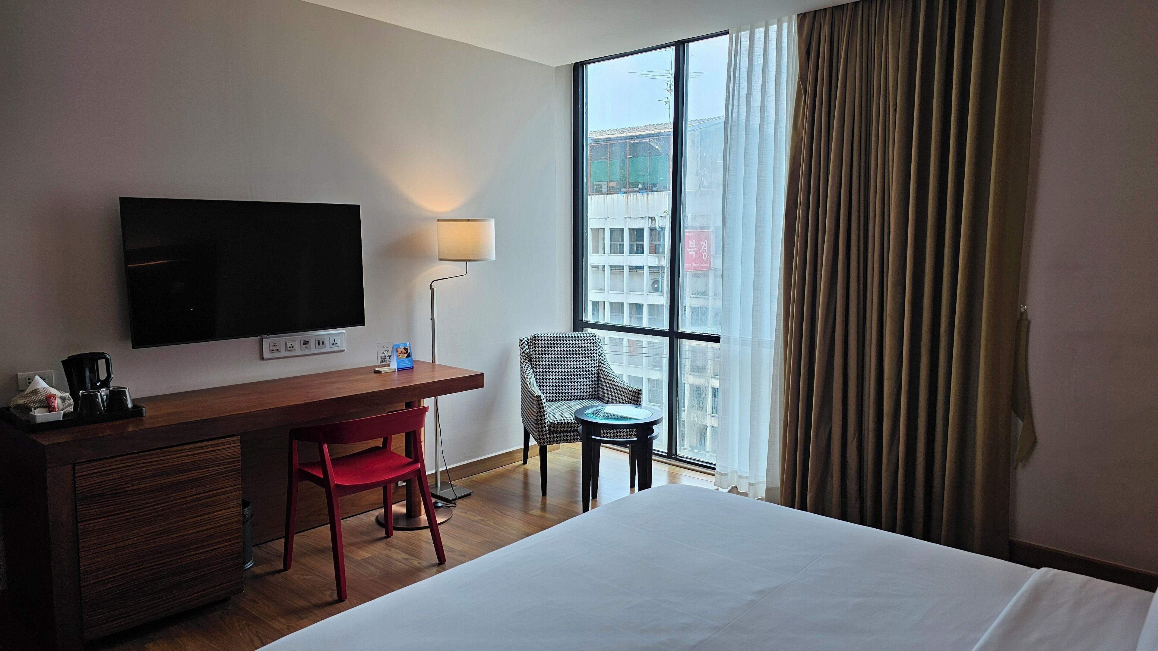 A hotel room with a bed, a desk with a TV, a chair, a small table, and a window with curtains.