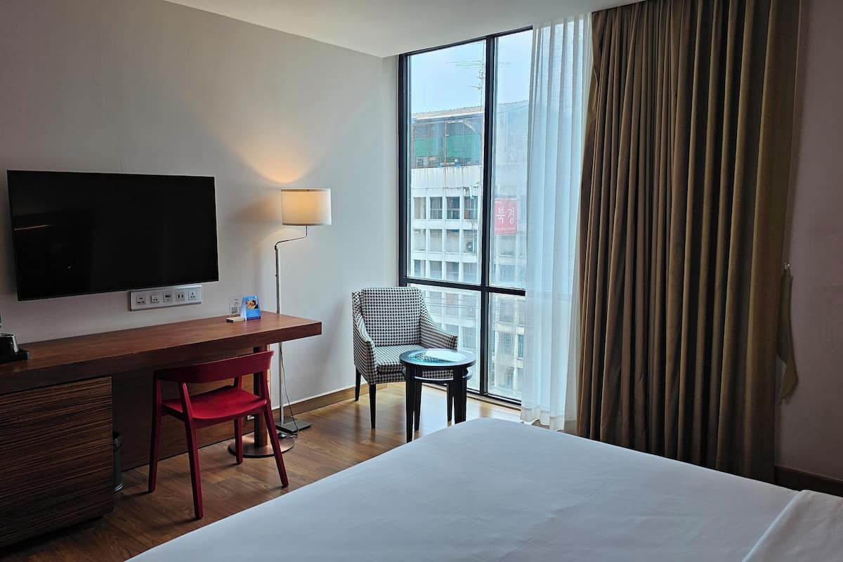 A hotel room with a bed, a desk with a TV, a chair, a small table, and a window with curtains.