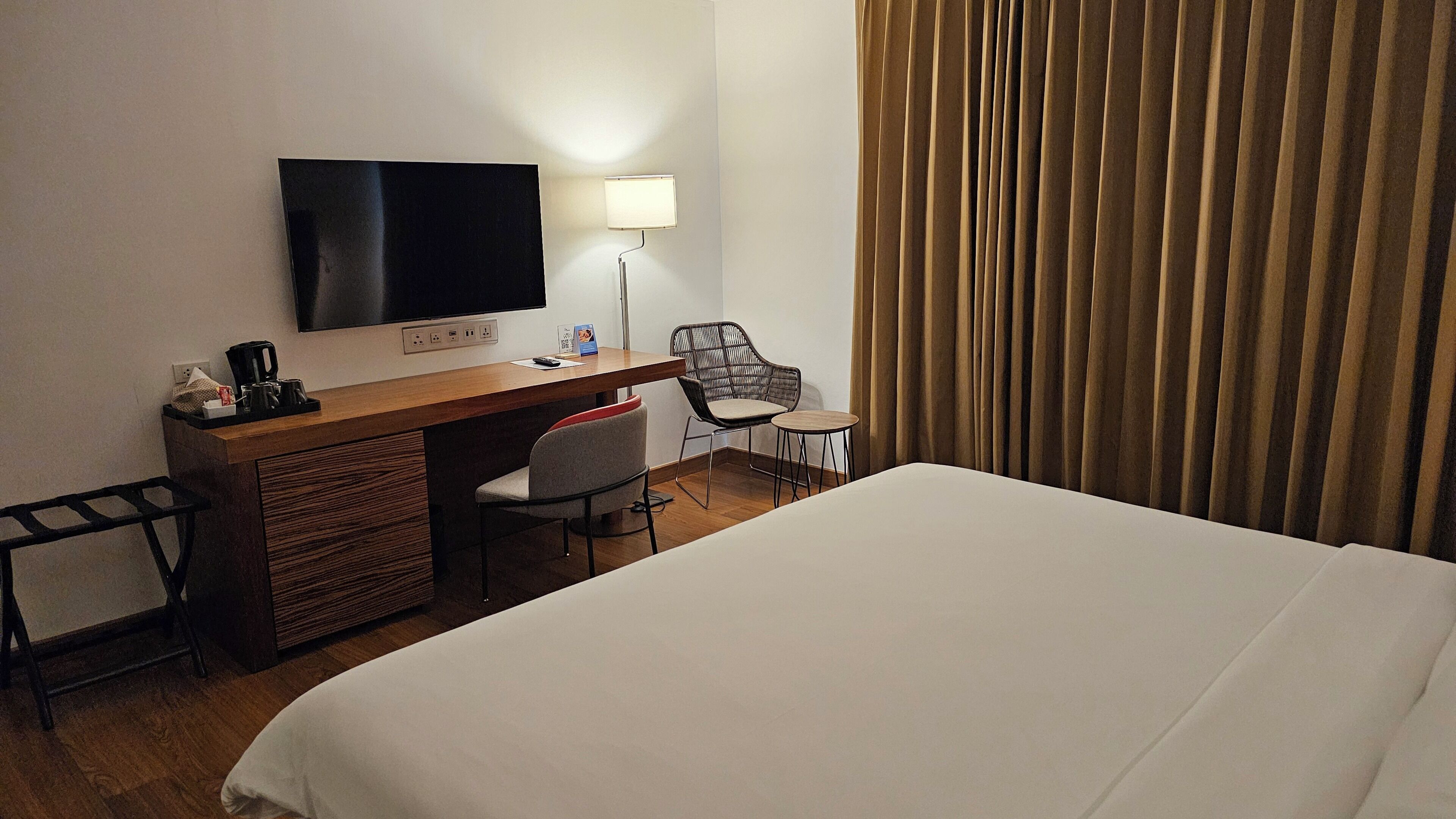 A hotel room with a bed, a desk with a flat-screen TV, a chair, a small table, and a window with curtains.