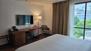 A hotel room with a bed, desk, chair, TV, lamp, and a window with a view of buildings and trees. - Sachas Hotel Uno Asoke Sukhumvit Bangkok (Bangkok)