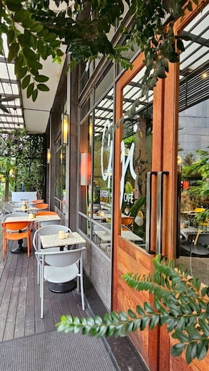 An outdoor cafe with tables and chairs, a wooden facade, and a glass window with a cafe sign. - Sachas Hotel Uno Asoke Sukhumvit Bangkok (Bangkok)
