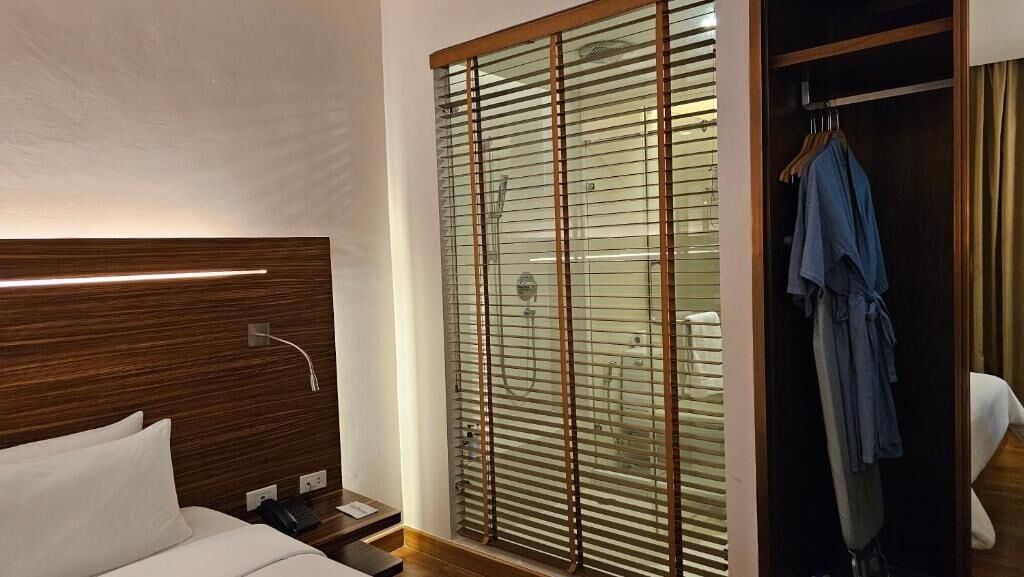 Quarto de hotel com cabeceira de madeira, mesa de cabeceira com telefone e um amplo guarda-roupa com porta de correr de vidro, onde se vê um roupão pendurado.