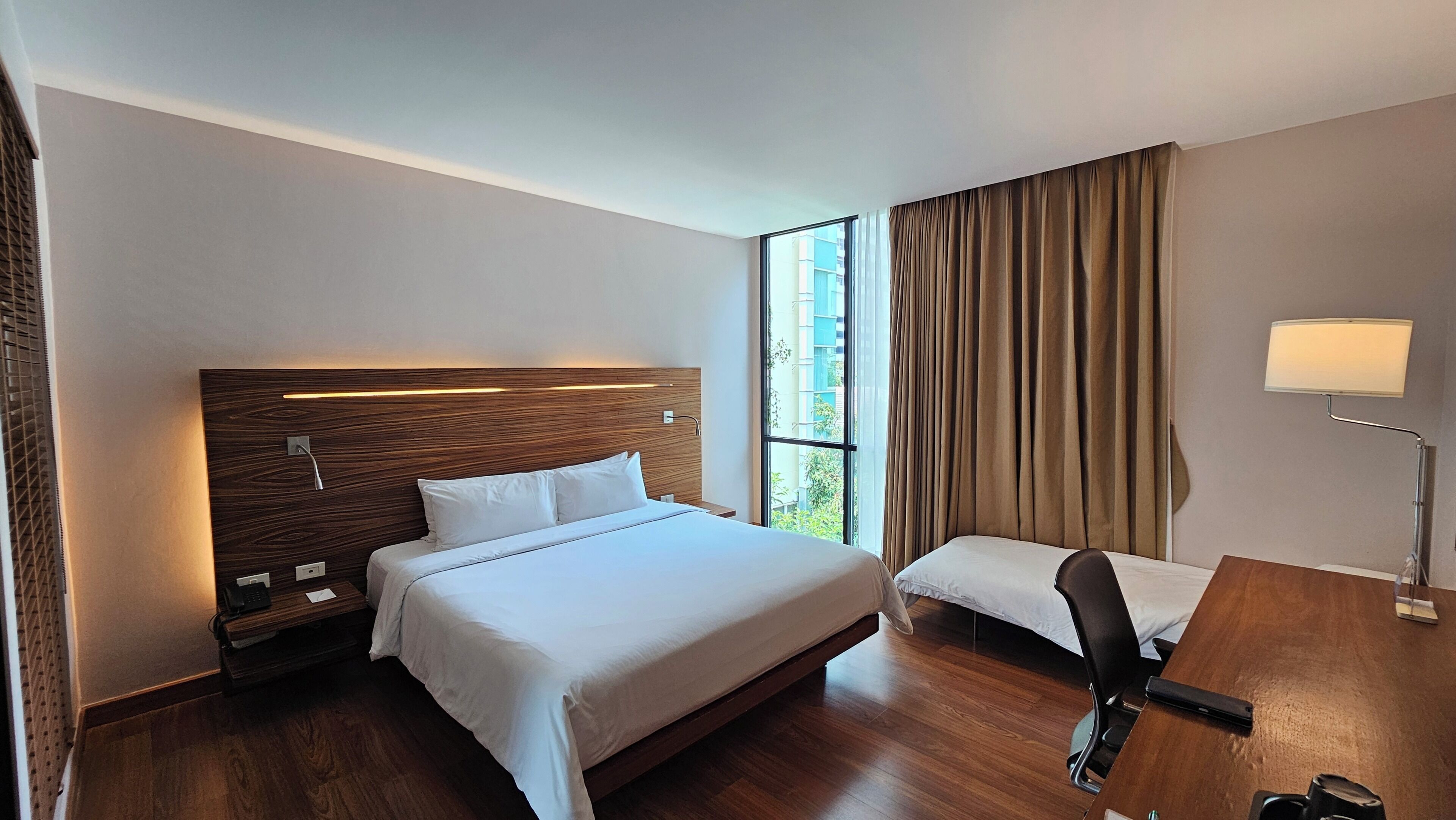 A hotel room with a large bed, a desk, a chair, a lamp, and a window with curtains.