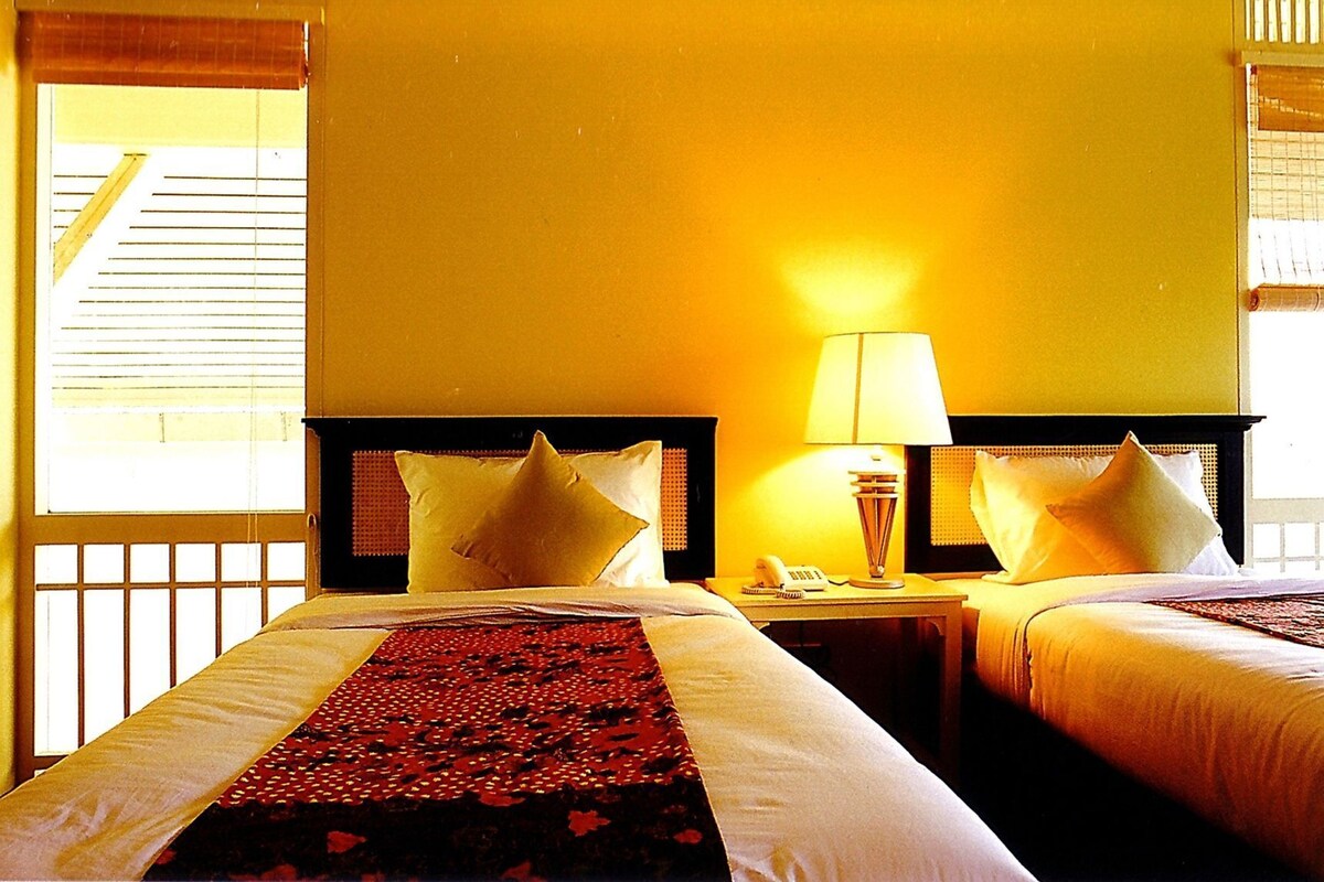 Quarto de hotel com duas camas, uma mesa de cabeceira com abajur e uma janela com persianas.