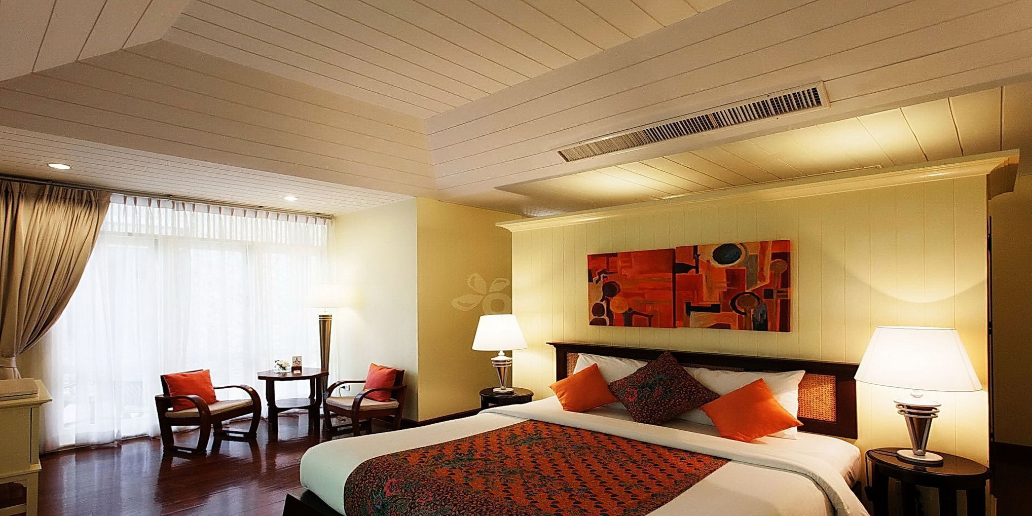 A hotel room with a bed, two bedside tables, a desk with chairs, a large abstract painting on the wall, and a window with curtains.